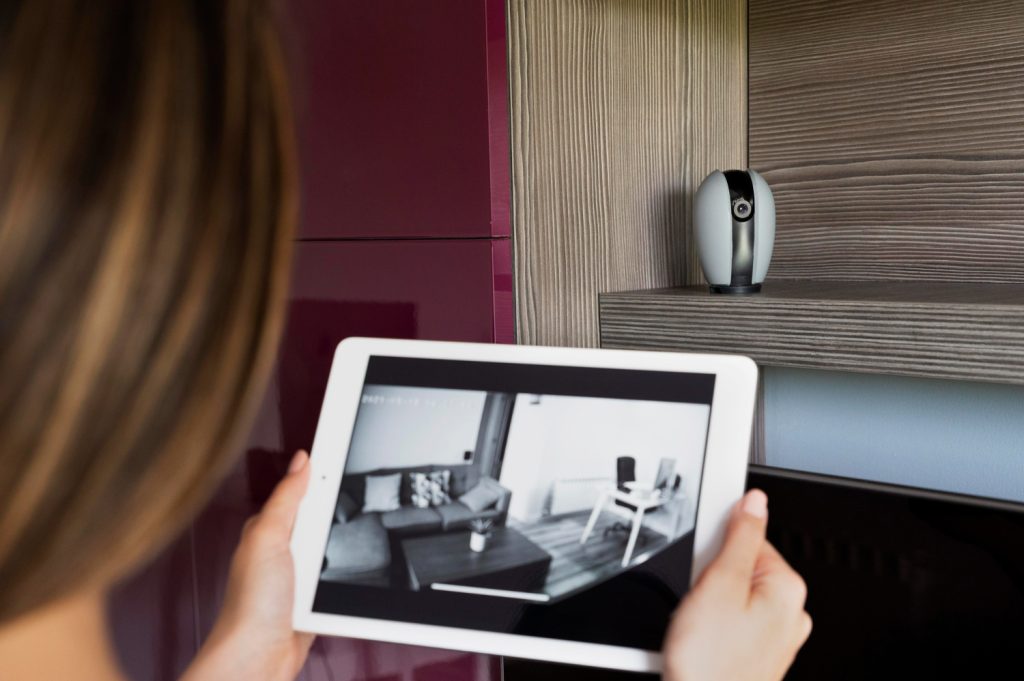 A person holding a tablet showing a live black-and-white feed from a small indoor Wi-Fi camera on a shelf.