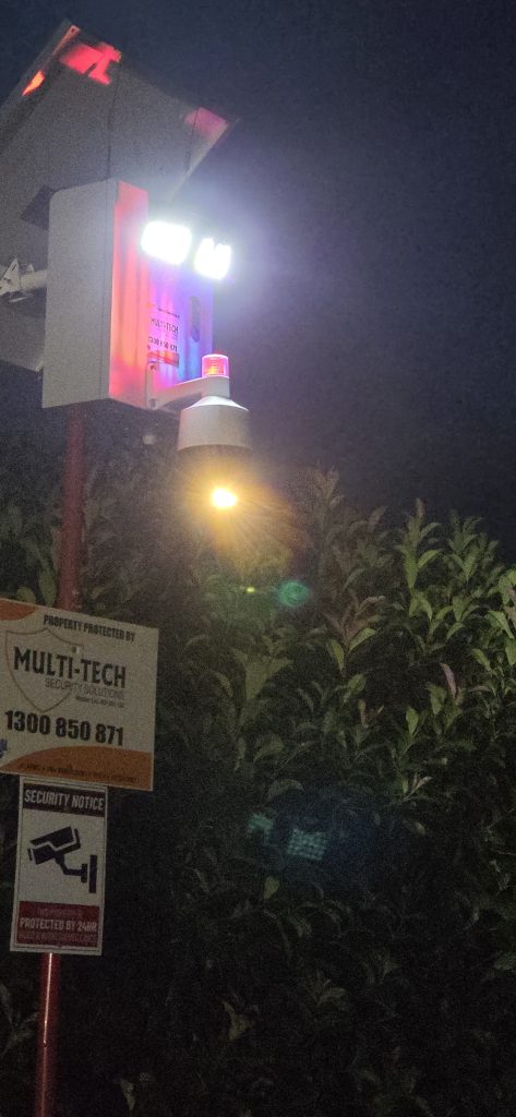 Multi-Tech security camera at night with active red and blue flashing lights and a bright floodlight.