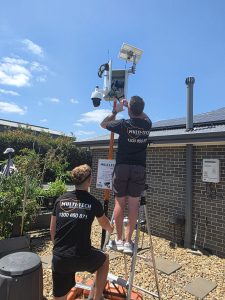 Two technicians from Multi-Tech Security Solutions installing a solar-powered CCTV camera system on a pole.