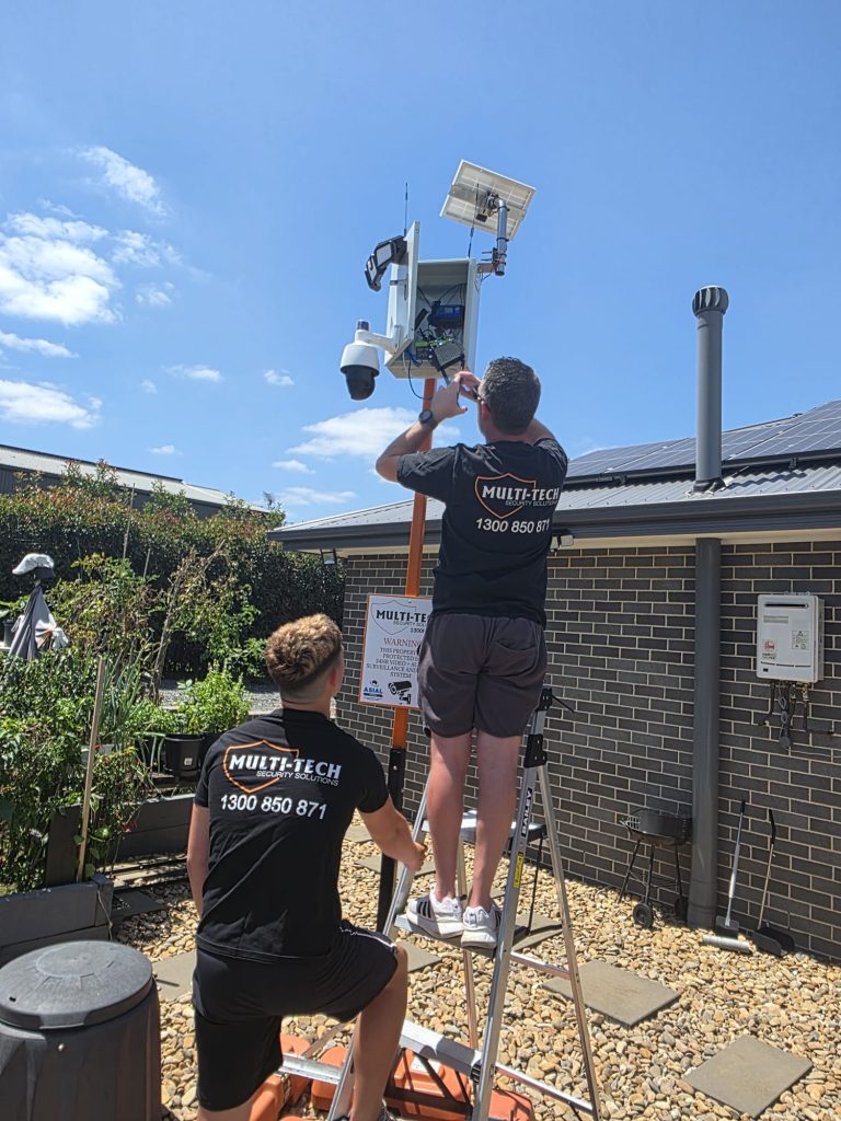 Two technicians from Multi-Tech Security Solutions installing a solar-powered CCTV camera system on a pole.